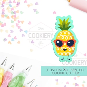 May include: A 3D printed cookie cutter shaped like a pineapple wearing sunglasses. The pineapple is yellow with a green top and red sunglasses. The text "CUSTOM 3D PRINTED COOKIE CUTTER" is on a light blue background.