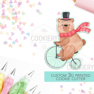 May include: Custom 3D printed cookie cutter featuring a brown bear wearing a red scarf and a top hat, riding a bicycle. The cutter is on a white background with colorful heart sprinkles.