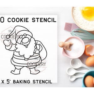 May include: A white 12.7cm x 12.7cm baking stencil featuring a Santa Claus design. The stencil reads "PYO COOKIE STENCIL" and has "The COOKIERY MAMA" logo. Baking ingredients, such as eggs, flour, and sugar, are in the background.