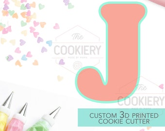 May include: A pink 3D printed cookie cutter in the shape of the letter J. The cutter is on a white background with sprinkles and piping bags.
