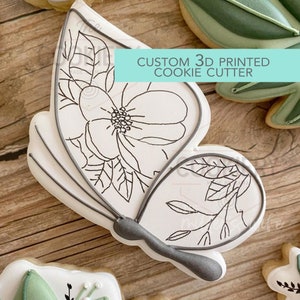 May include: A white cookie with a black outline of a butterfly with a flower and leaves. The cookie is on a wooden surface. The text "CUSTOM 3D PRINTED COOKIE CUTTER" is in the upper right corner of the image.