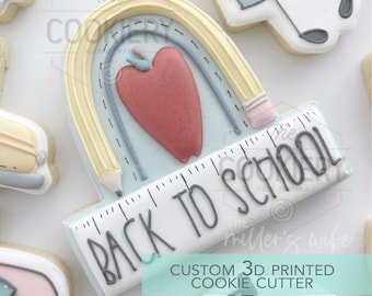 Op de afbeelding: Een blauwe en witte koekjesvorm in de vorm van een liniaal met de tekst "Back to School" en een ontwerp van een gele potlood en een rode appel. De koekjesvorm is een aangepast 3D-geprint ontwerp.