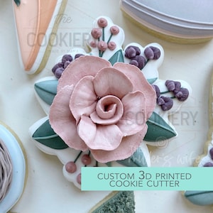 Può includere: Un tagliapasta stampato in 3D a forma di rosa rosa con foglie verdi e bacche viola. Il testo "CUSTOM 3D PRINTED COOKIE CUTTER" è su uno sfondo turchese.