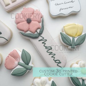 Puede incluir: Surtido de galletas decoradas con diseños florales. Una galleta presenta una flor rosa con la palabra "mama" escrita en verde. Otras galletas incluyen un tulipán amarillo y un mensaje que dice "their hearts forever". El texto "Custom 3D Printed Cookie Cutter" también es visible.