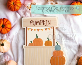 May include: A 3D printed cookie cutter shaped like a pumpkin stand. The stand has a sign that says "Pumpkin" and a banner with flags. There are four pumpkins on the stand and a stack of three pumpkins on the right side.