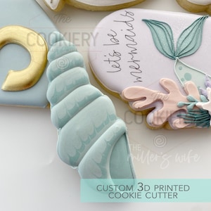 Pode incluir: Um cortador de biscoitos impresso em 3D com a forma de uma concha. O cortador é azul claro com uma superfície texturizada. O texto "Let's be mermaids" é visível no fundo.
