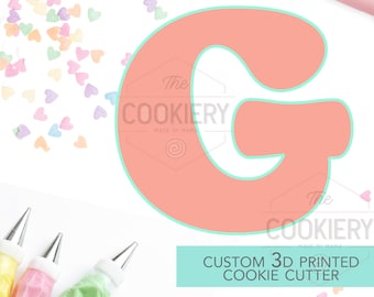 May include: A light pink 3D printed cookie cutter in the shape of the letter G. The cutter is outlined in a light turquoise color. The cutter is surrounded by colorful heart sprinkles and piping bags. The text "Custom 3D Printed Cookie Cutter" is displayed in white on a turquoise background.