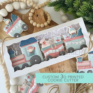 Puede incluir: Una caja blanca con una ventana transparente que muestra un conjunto de cuatro cortadores de galletas de Navidad con forma de tren. El tren está decorado con glaseado azul, rojo y blanco y tiene las palabras "la la la la la la" escritas en él. La caja está etiquetada como "Cortador de galletas personalizado 3D impreso".