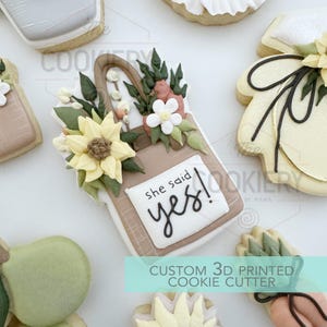 Może przedstawiać: Ozdobione ciasteczka maślane z motywami kwiatowymi i torebkami. Jedno ciastko przedstawia torebkę ze słonecznikiem i napisem "she said yes!". Inne ciastka mają wzory gruszek, sukienek i inne kwiatowe. Obraz zawiera również tekst "CUSTOM 3D PRINTED COOKIE CUTTER".