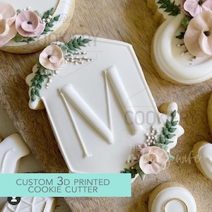 May include: Custom 3D printed cookie cutter in the shape of a house with the letter M in the centre. The cutter is decorated with white icing and green leaves. The text "CUSTOM 3D PRINTED COOKIE CUTTER" is visible at the bottom of the image.