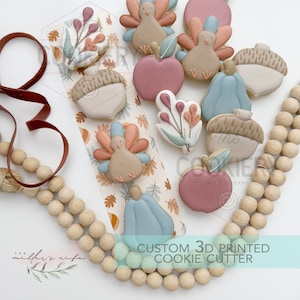 May include: A collection of autumn-themed cookies shaped like turkeys, pumpkins, acorns, apples, and floral sprigs, arranged on a patterned card. The set includes a dark red ribbon and a string of natural wooden beads, with text reading CUSTOM 3D PRINTED COOKIE CUTTER.
