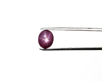 May include: A deep red star sapphire gemstone held in tweezers. The gemstone has a star-like pattern in the center.
