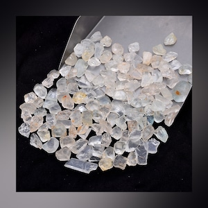 May include: A large pile of clear, faceted, aquamarine gemstones. The stones are various sizes and shapes, and they are scattered on a silver metal surface.