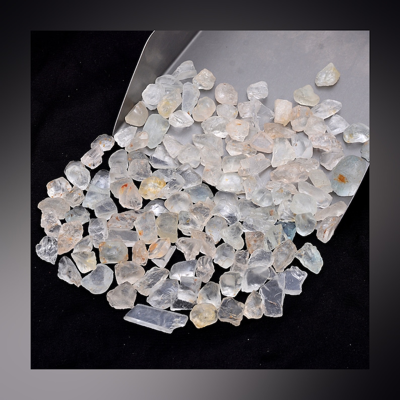 May include: A large pile of clear, faceted, aquamarine gemstones. The stones are various sizes and shapes, and they are scattered on a silver metal surface.