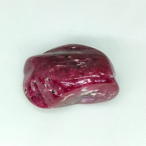 Puede incluir: Una piedra preciosa pulida, de color rojo intenso, con inclusiones blancas y negras. La piedra lisa y redondeada tiene una superficie brillante y una forma natural. La piedra preciosa está sobre un fondo blanco.