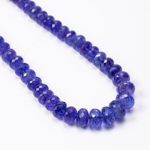 May include: A necklace of faceted, deep blue gemstones. The beads are strung together, creating a curved line against a white background. The gemstones have a sparkling, polished appearance, reflecting light.