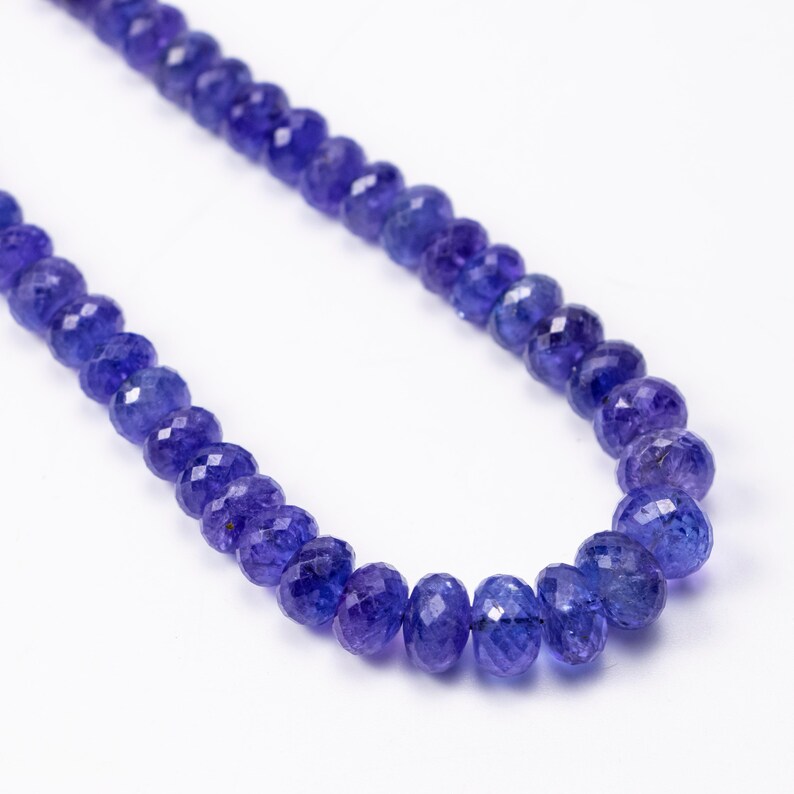 May include: A necklace of faceted, deep blue gemstones. The beads are strung together, creating a curved line against a white background. The gemstones have a sparkling, polished appearance, reflecting light.