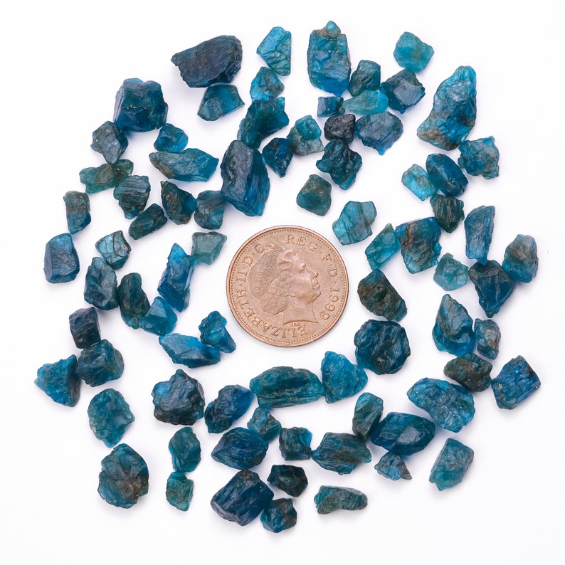 May include: A collection of blue-green gemstone chips, scattered around a British one pound coin. The coin features a portrait of Queen Elizabeth II.