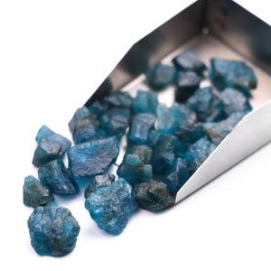 May include: A metal scoop filled with loose blue gemstones. The gemstones are various sizes and shapes, and some are spilling out of the scoop onto a white surface.