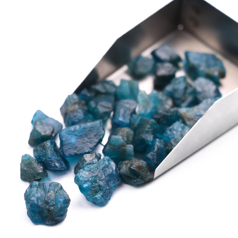 May include: A metal scoop filled with loose blue gemstones. The gemstones are various sizes and shapes, and some are spilling out of the scoop onto a white surface.