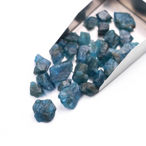 May include: A close-up of a metal scoop filled with a pile of blue gemstones. The gemstones are scattered on a white surface.
