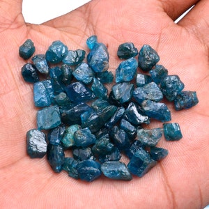 May include: A pile of blue apatite rough stones, also known as blue apatite crystals, on a hand. The stones are various sizes and shapes, with some having a more pointed shape.