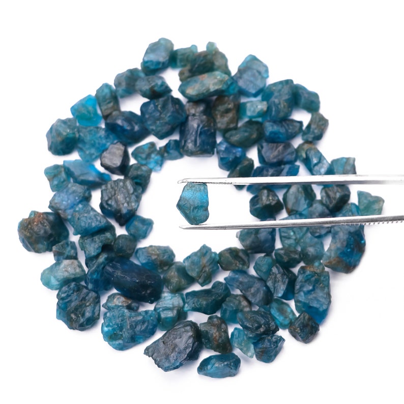 May include: A collection of blue-green apatite crystals, some of which are being held by a pair of tweezers. The crystals are various sizes and shapes, and have a glassy luster.