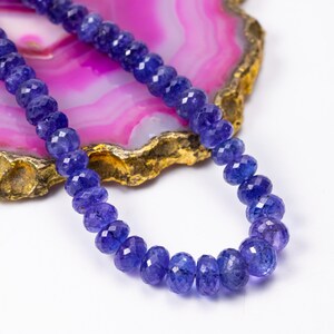 May include: A necklace of faceted, deep blue gemstones. The beads are strung together, creating a continuous loop. The background features a pink and gold-edged stone slice, providing a contrasting backdrop for the jewelry.