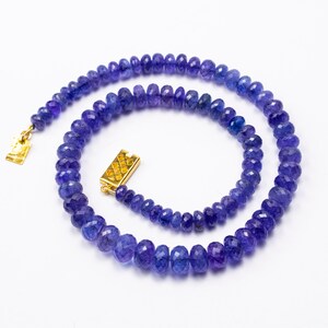 May include: A necklace composed of faceted, deep blue-violet gemstones. The beads are strung together, forming a circular shape. The necklace features a gold-colored clasp. The gemstones have a polished, reflective surface, and the necklace is set against a white background.