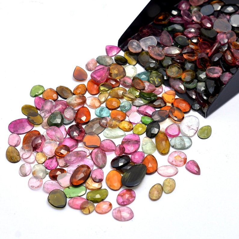 May include: A large assortment of colorful, faceted gemstones in various shades of pink, green, brown, and black. The gemstones are loose and scattered on a white surface, with some spilling out of a black container.