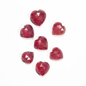 Op de afbeelding: Zeven rode hartvormige edelstenen met gefacetteerde oppervlakken. De edelstenen zijn verspreid op een witte achtergrond gerangschikt.