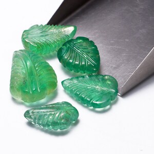 May include: Six green gemstone leaf-shaped beads, carved with intricate details, are scattered on a white surface. The beads are spilling out of a metal scoop.