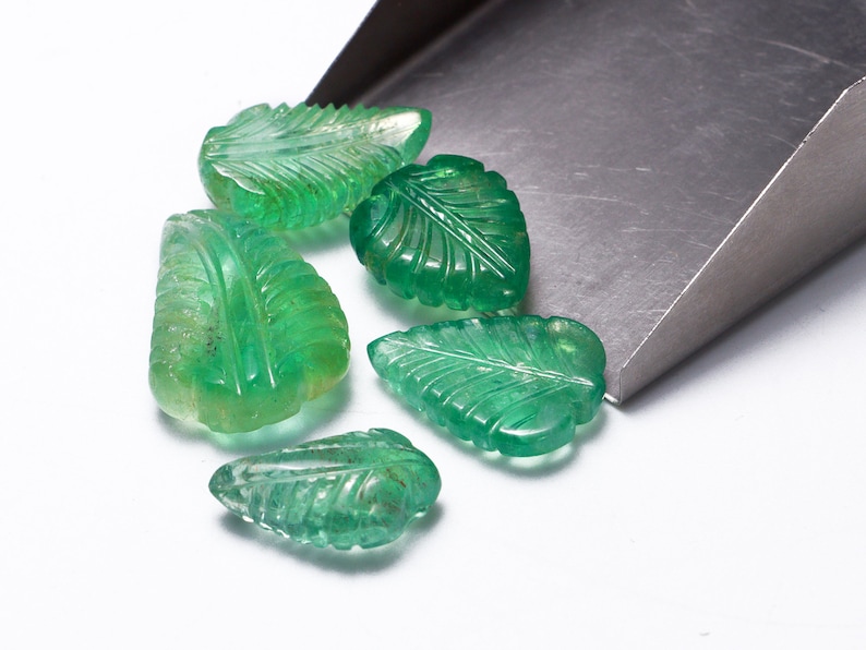 May include: Six green gemstone leaf-shaped beads, carved with intricate details, are scattered on a white surface. The beads are spilling out of a metal scoop.