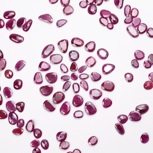 May include: A collection of loose, faceted, pink ruby gemstones. The rubies are various sizes and shapes, with some having a more rounded, oval shape, and others having a more elongated, rectangular shape.