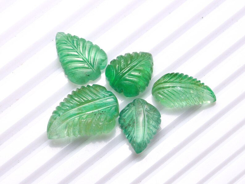 May include: Five green gemstone leaf-shaped beads. The beads are carved with detailed veins and are a light to medium shade of green.