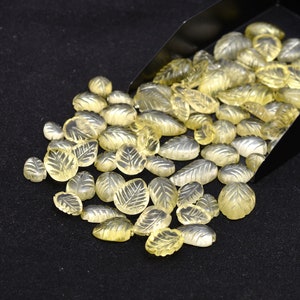 May include: A large group of translucent yellow leaf-shaped gemstones, some with intricate carvings, are scattered on a black surface. The gemstones appear to be spilling out of a black metal scoop.