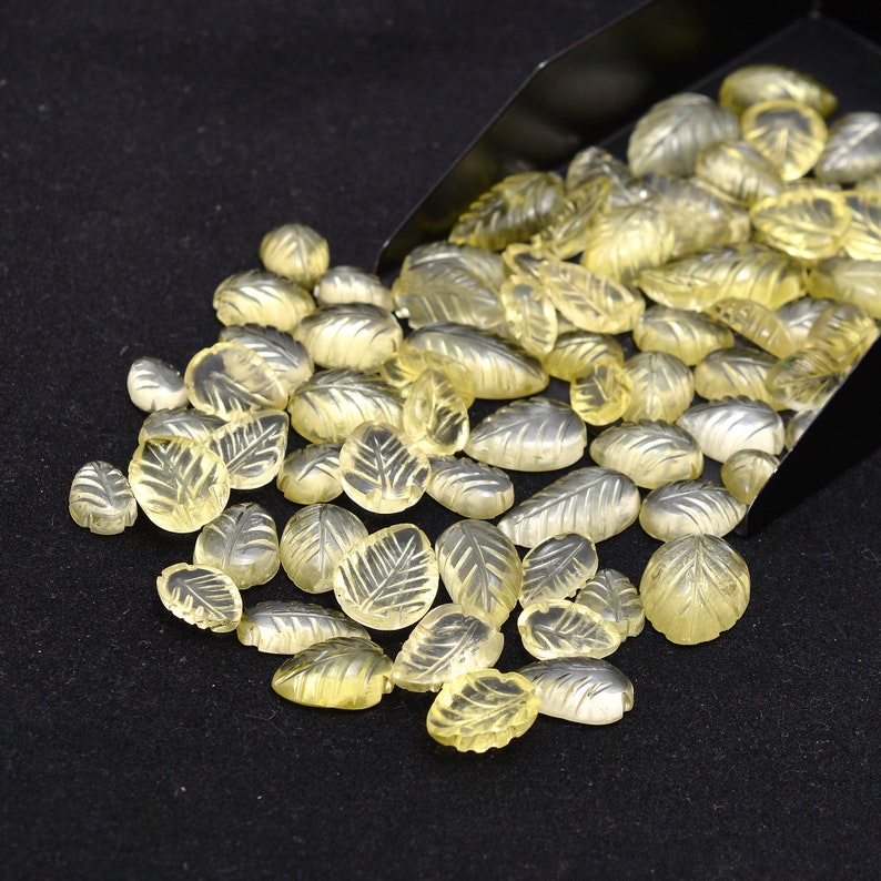 May include: A large group of translucent yellow leaf-shaped gemstones, some with intricate carvings, are scattered on a black surface. The gemstones appear to be spilling out of a black metal scoop.