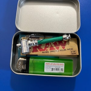 May include: A silver metal tin containing a small metal pipe with a green handle, a pack of RAW natural unrefined rolling papers, and a green lighter. The rolling papers are 1 1/4 size.