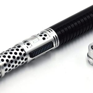Puede incluir: Un sable de luz plateado y negro con un cañón de metal perforado y un mango negro con un agarre texturizado. El sable de luz está desmontado, mostrando las partes separadas.