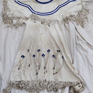 May include: A white leather garment with fringe detailing and blue trim around the neckline. The garment has a decorative design of blue and white beads around the neckline.
