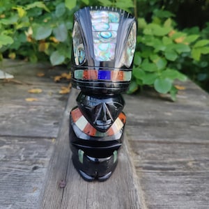 Op de afbeelding: Een zwart stenen beeldje met een gestileerd hoofd en een inleg van iriserend abalone-schelpmateriaal op de kroon. Het figuur bevat een band van kleurrijke stenen inleg in blauw, rood en groen, met extra stenen accenten rond de nek en de basis.