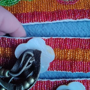May include: Close-up of a beaded textile with a floral design. The textile features red, orange, and gold beads, with a blue felt section. White, flower-shaped buttons with leather straps and gold beads are attached.