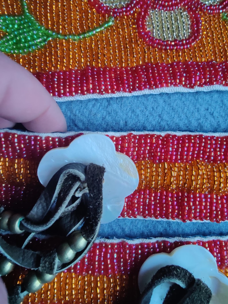 May include: Close-up of a beaded textile with a floral design. The textile features red, orange, and gold beads, with a blue felt section. White, flower-shaped buttons with leather straps and gold beads are attached.