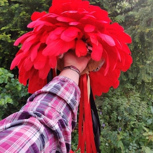 Puede incluir: Un tocado de plumas rojas con cintas negras y rojas que cuelgan de la parte inferior. El tocado está sostenido por una mano que lleva una pulsera de plata.