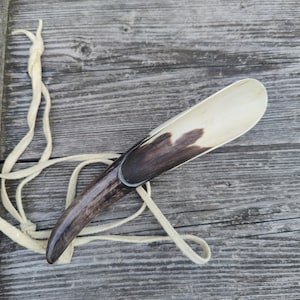 Puede incluir: Un calzador artesanal con un diseño curvo y alargado. El calzador presenta un mango marrón oscuro que hace la transición a un color de madera más claro en la pala. Se adjunta un cordón de cuero color crema para colgar.