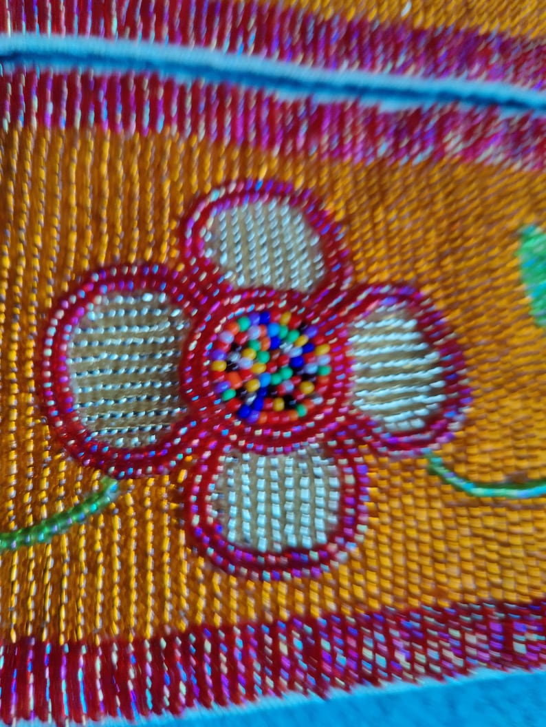 May include: Close-up of a beaded textile with a floral design. The flower is red with a center of multicolored beads. The background is orange with vertical beadwork, and the edges are trimmed in red.