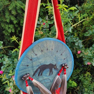 Peut inclure: Un tambour rond bleu peint à la main avec une illustration de loup noir et des formes en forme de spermatozoïdes. Le tambour a une sangle rouge et crème et est orné de plumes et de cloches. Le fond est une verdure luxuriante.