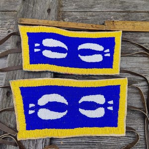 May include: Two beaded rectangular panels with a blue background and white curved shapes. The panels have a yellow border and brown leather straps. The panels are displayed on a weathered wooden surface.