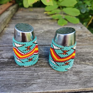 Pode incluir: Dois saleiros e pimenteiros com contas. Os saleiros e pimenteiros têm uma parte superior prateada e são cobertos por contas turquesas com um padrão colorido de vermelho, amarelo e branco. Estão sobre uma superfície de madeira envelhecida.