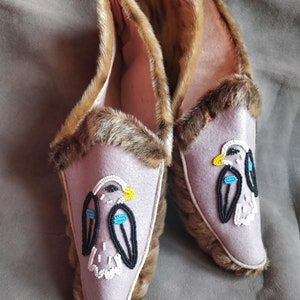 Puede incluir: Un par de zapatillas de piel marrón con ribete de piel y un interior de tela de color morado claro. Cada zapatilla presenta un diseño de cuentas que representa un pájaro con cuentas negras, blancas, azules y amarillas.